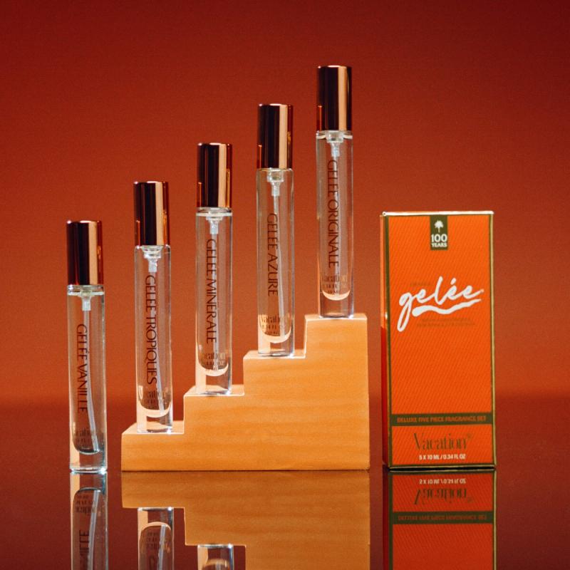 Five clear perfume rollerball bottles with copper caps on a tiered wooden display, next to an orange "Gelée" perfume box, against an orange background.