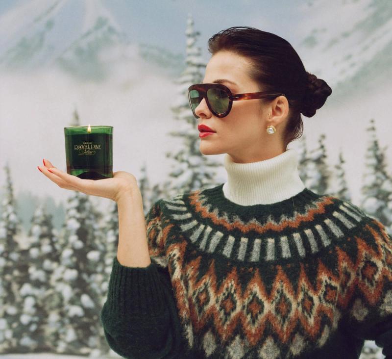 a woman in a sweater and sunglasses is holding a Royal Pine candle in her hand 