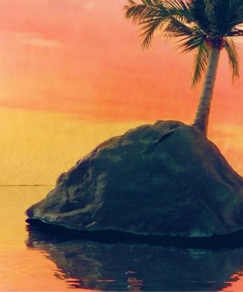 A small rocky island with a single palm tree stands in calm water under a vibrant orange and yellow sunset.