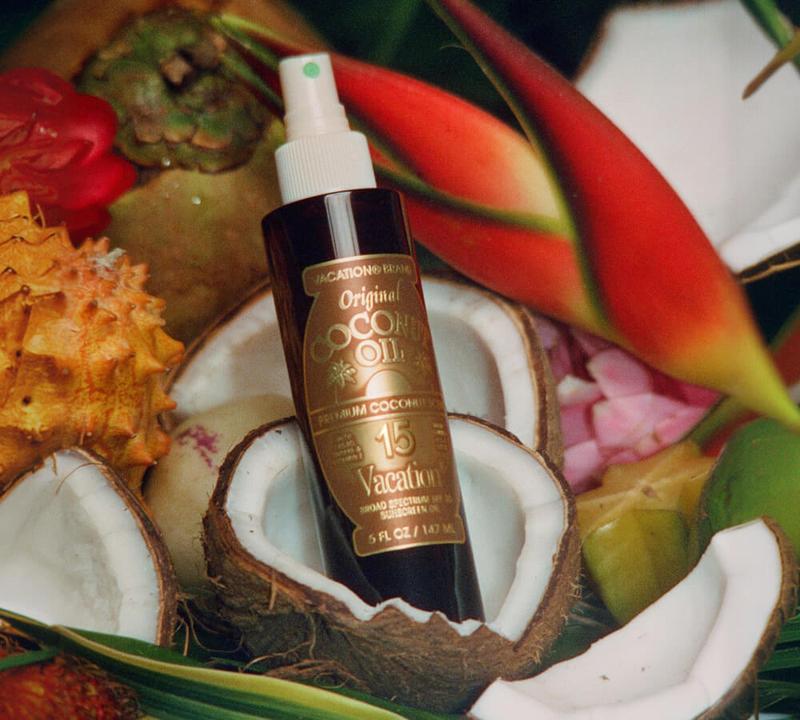 A bottle of Vacation Brand Original Coconut Oil with SPF 15, surrounded by coconut halves, tropical fruits, and red flowers.
