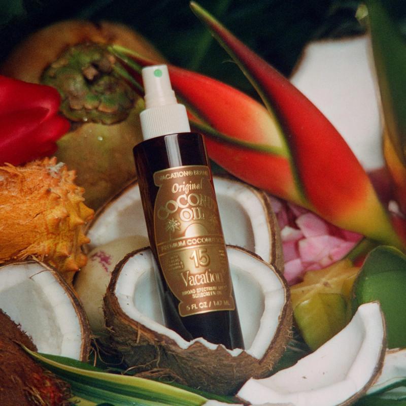 A dark brown spray bottle of Vacation Original Coconut Oil SPF 15 sunscreen is nestled among split coconuts, tropical fruits, and red flowers.