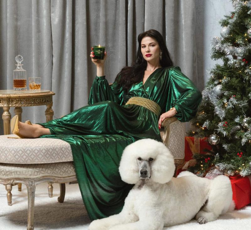 a woman in a green satin dress is holding a candle and sitting on a chair next to a white poodle named Roman