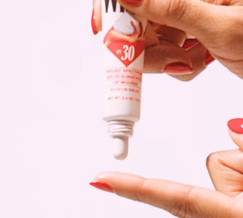 A hand with red nail polish squeezes SPF 30 lip balm from a tube onto a fingertip.