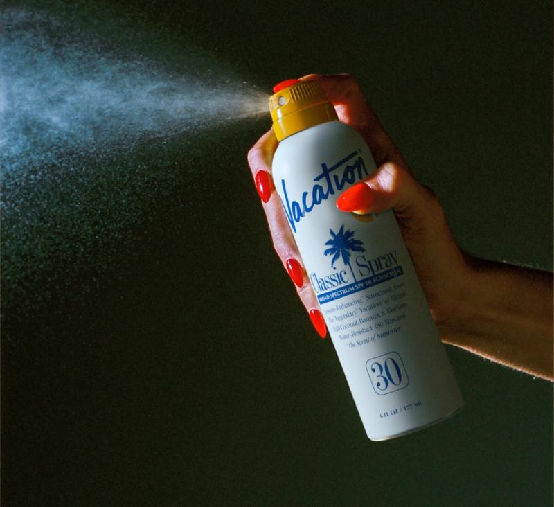 Hand with red nails spraying "Vacation Classic Spray" sunscreen, creating a visible mist.