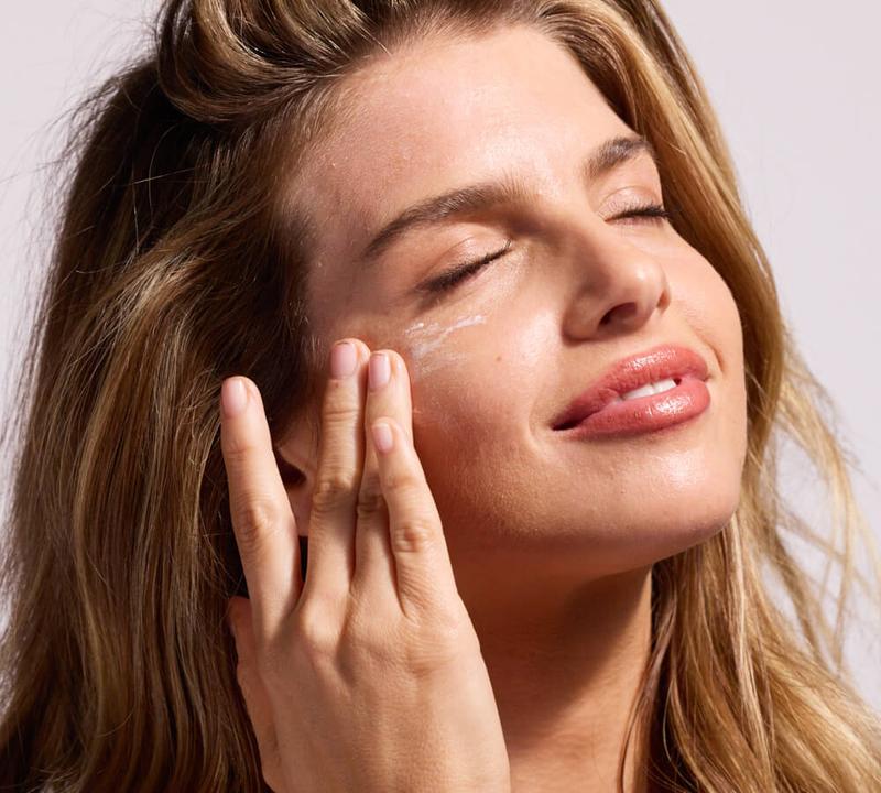 A woman with closed eyes applies cream to her cheek.