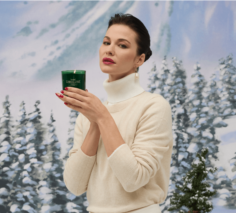 a woman in a beige cashmere sweater holding a green candle on a snowy alpine backdrop