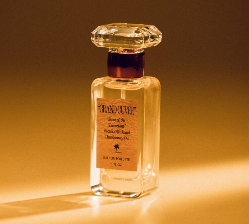 A perfume bottle with a clear, rectangular body and faceted cap, labeled "GRAND CUVÉE Scent of the 'Luxuriant' Vacation® Brand Chardonnay Oil Eau de Toilette" on a golden background.
