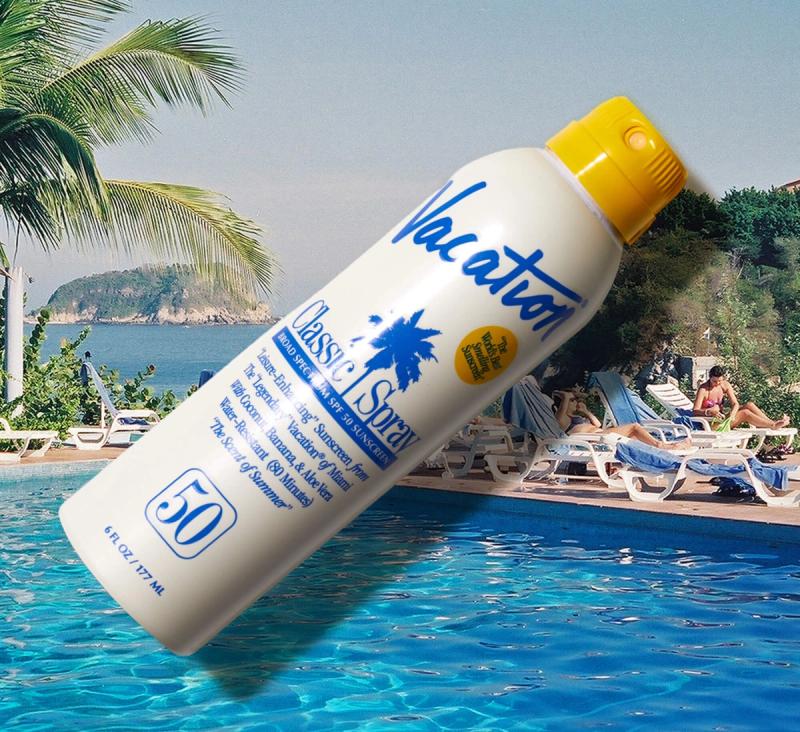 A white Vacation Classic Spray SPF 50 sunscreen bottle with a yellow cap floats near a swimming pool with a tropical beach resort in the background.