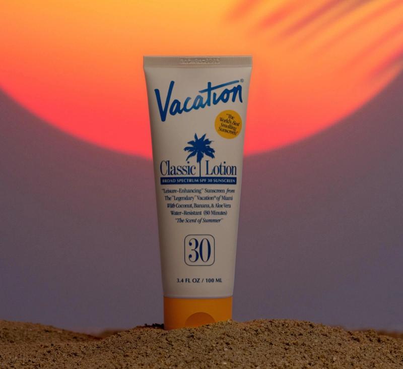 A tube of Vacation Classic Lotion SPF 30 Sunscreen stands in sand against a sunset with a palm tree silhouette.