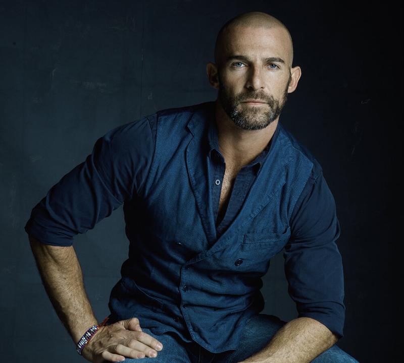 A bald, bearded man in a dark blue shirt and vest looks directly at the camera.