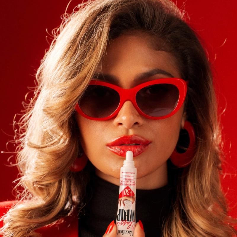 Woman in red sunglasses and lipstick holds Vacation Classic Whip SPF 30 to her lips on a red background.