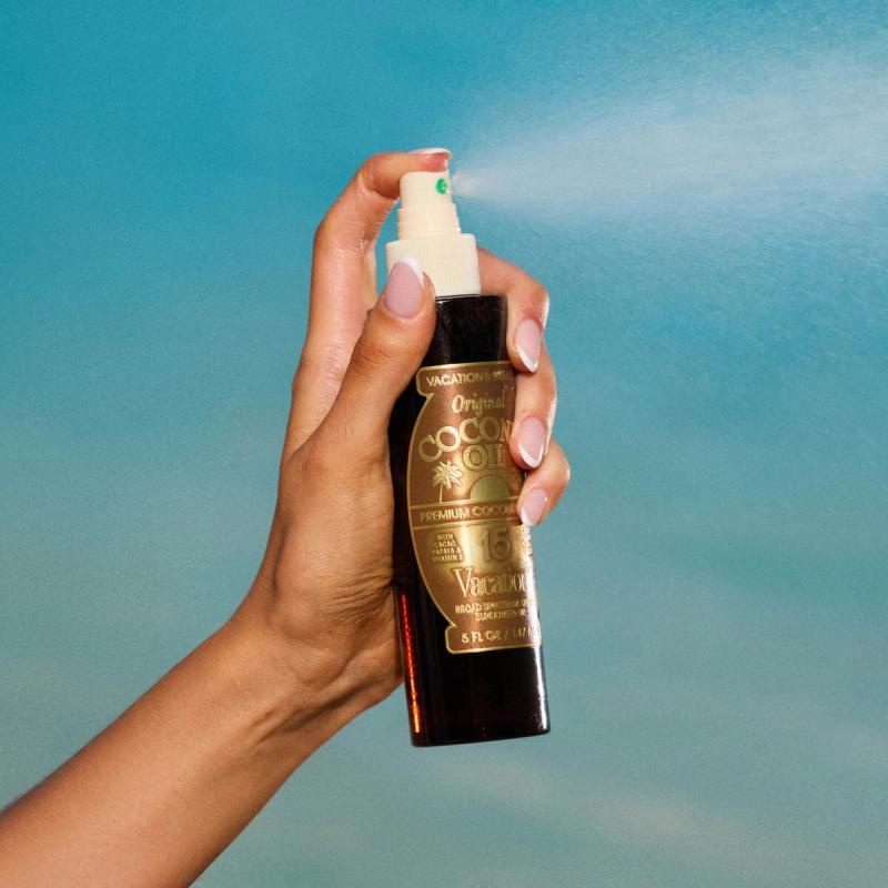 A hand sprays a bottle of Vacation Inc. Original Coconut Oil SPF 15, creating a fine mist against a blue background.