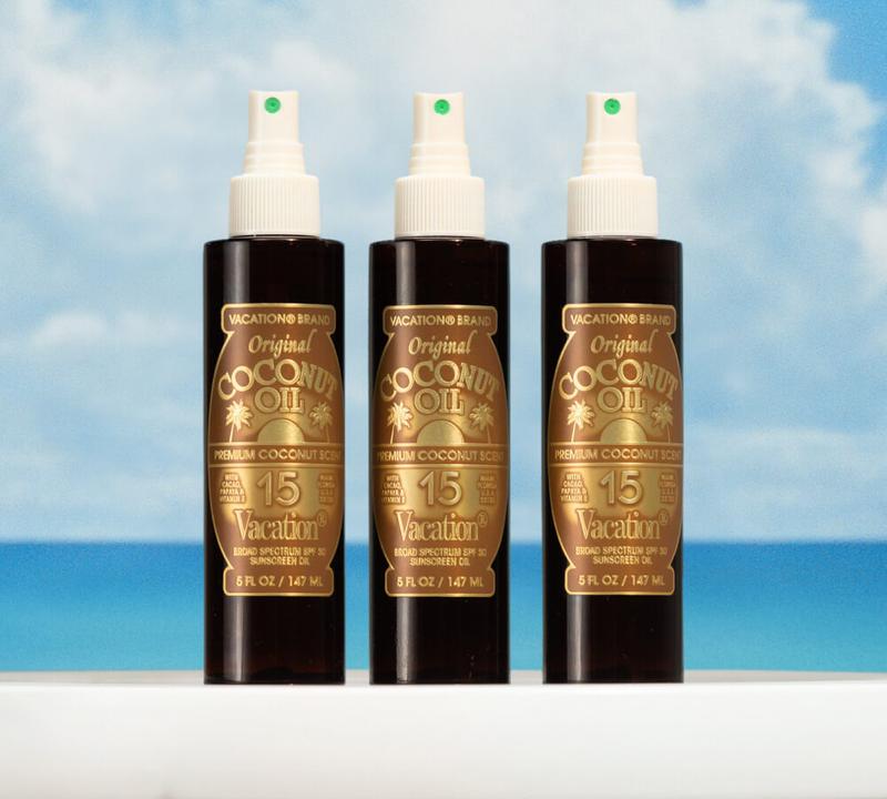 Three Vacation Brand Original Coconut Oil SPF 15 sunscreen spray bottles against a blue sky and ocean.