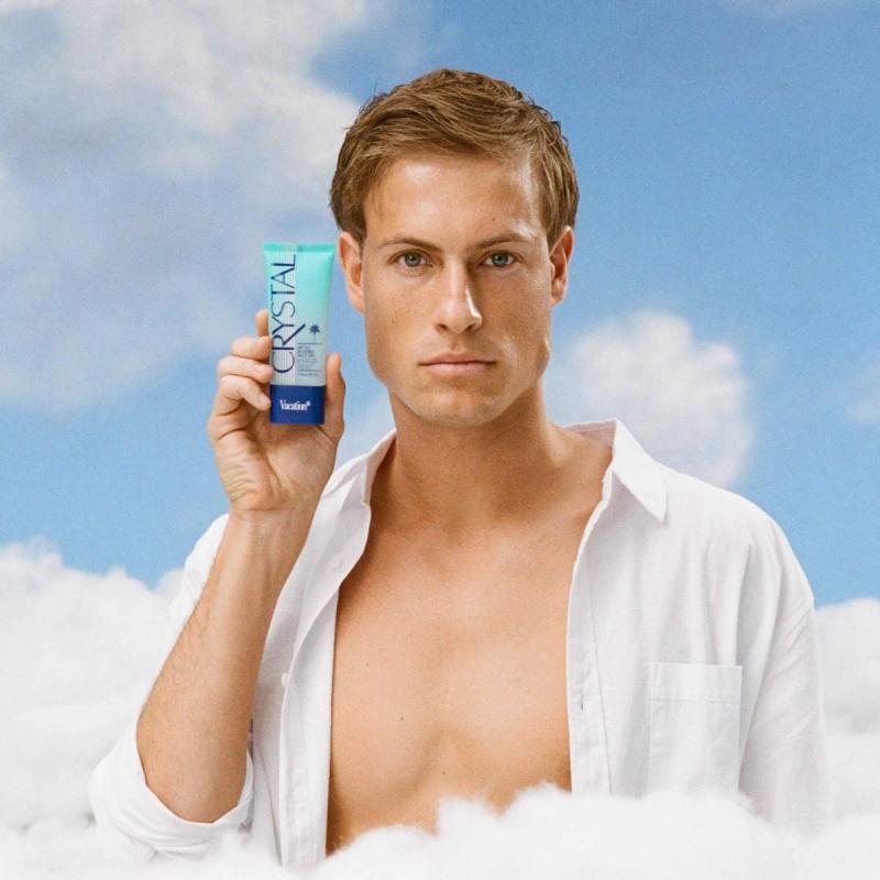 a man with an unbuttoned shirt is holding a bottle of Vacation® Crystal SPF 50 Invisible Face Gel