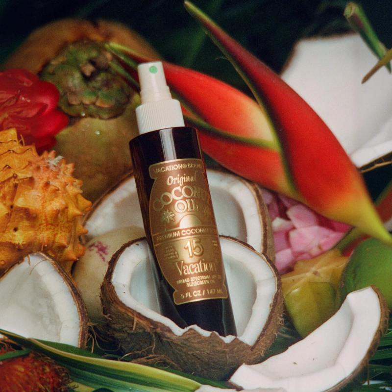 A bottle of Vacation Original Coconut Oil SPF 15 sunscreen surrounded by halved coconuts, tropical fruits, and red flowers.