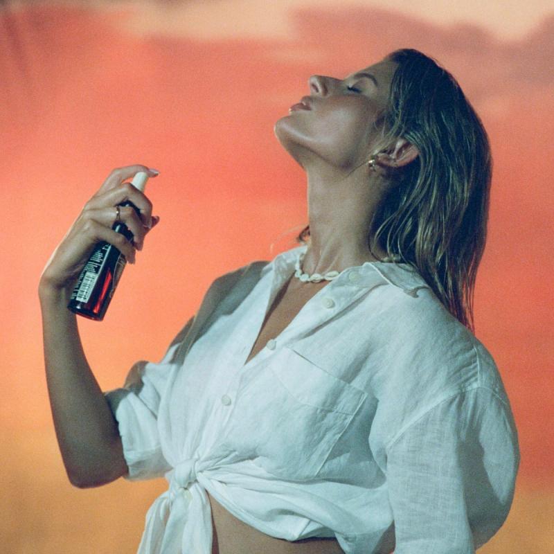 Profile of a woman with head tilted back, eyes closed, holding a spray bottle against a warm orange background.