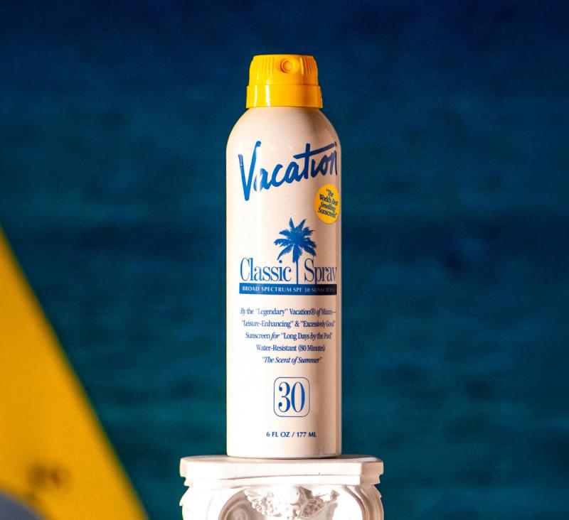 A white Vacation Classic Spray SPF 30 sunscreen bottle with a yellow cap, against a blurry blue background.