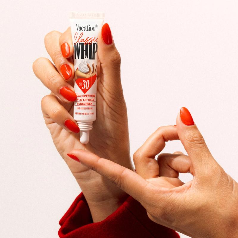 Hands with bright orange nails hold a tube of Vacation Classic WHIP SPF 30 Lip Balm Sunscreen.