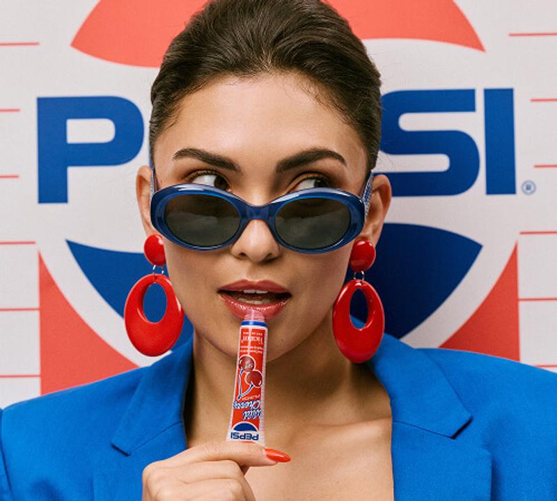 Woman in blue sunglasses and a blue blazer, holding a Pepsi Wild Cherry lip balm to her lips with a Pepsi logo in the background