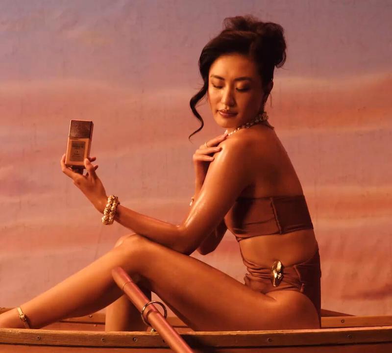 An Asian woman in a brown swimsuit and pearl jewelry sits in a boat, holding a small gold bottle, against a pastel sky.