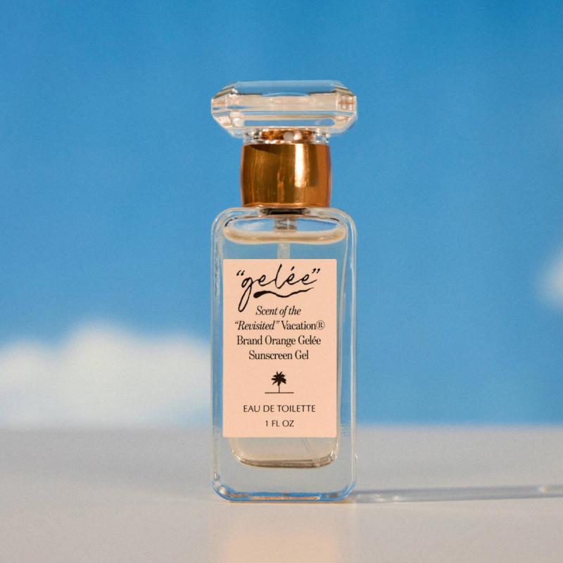 A clear glass "gélée" perfume bottle by Vacation® Brand with a peach label, copper cap, and faceted stopper, against a blue sky.