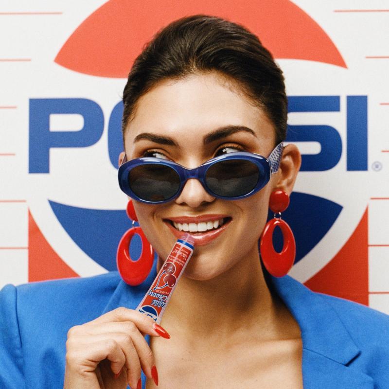 A woman in blue sunglasses and blazer holds Pepsi® Wild Cherry lip gel to her mouth, with a Pepsi logo in the background.