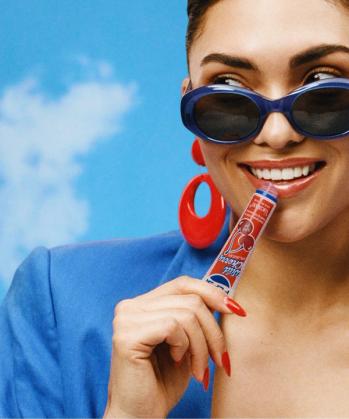 A smiling woman in blue sunglasses and red earrings applies a Wild Cherry Pepsi lip balm.
