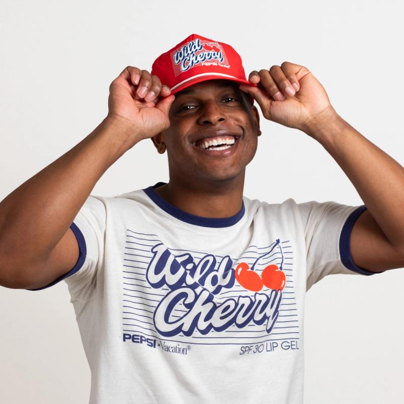 A smiling man wears a red "Wild Cherry Pepsi" hat and a white "Wild Cherry Pepsi" t-shirt.