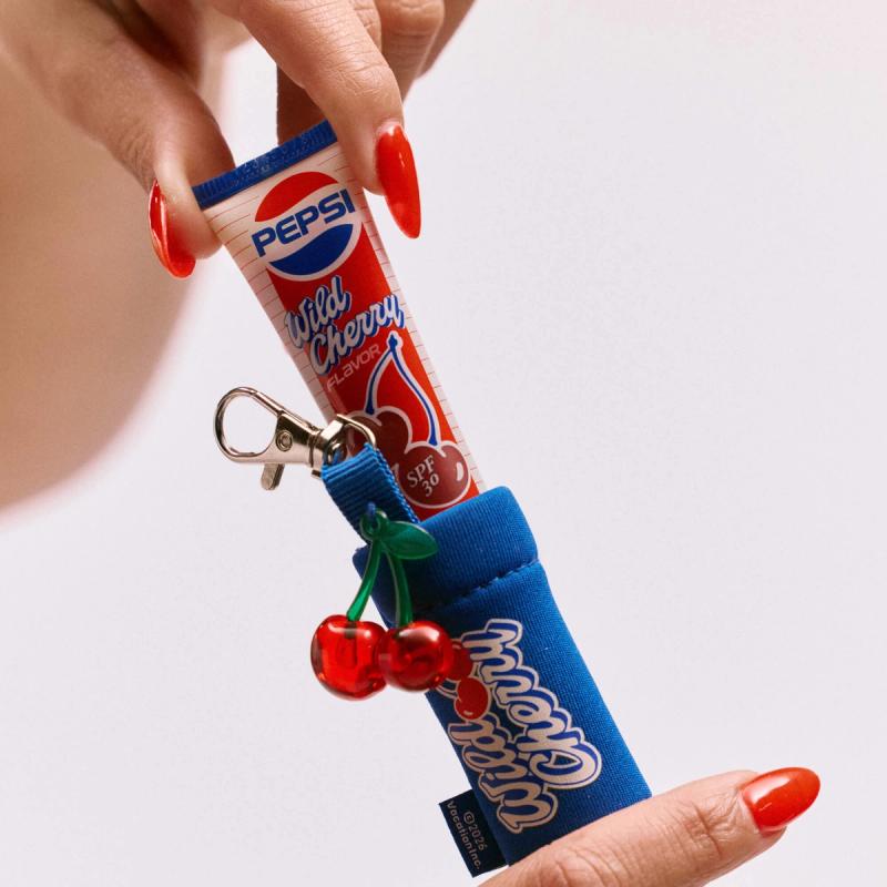 Person's hands with red nails holding a Pepsi Wild Cherry SPF 30 lip balm tube and a blue "Wild Cherry" holder with a cherry charm.