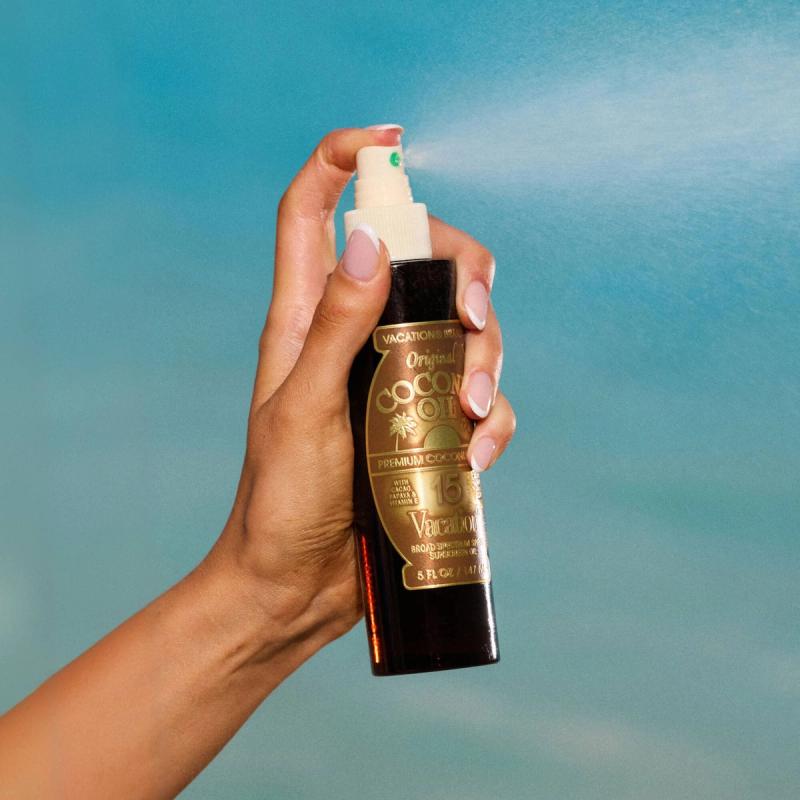 A hand sprays Vacation Inc. Original Coconut Oil, with a visible mist against a blue background.