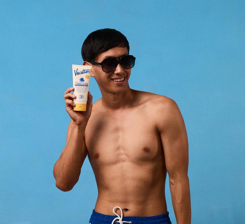 A smiling shirtless man in sunglasses holds up a tube of "Vacation" sunscreen against a blue background.