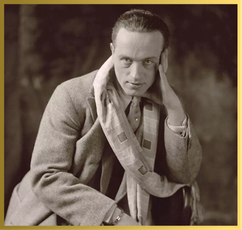 Sepia-toned portrait of a man in a tweed jacket and patterned scarf, hand to his temple, looking directly forward.