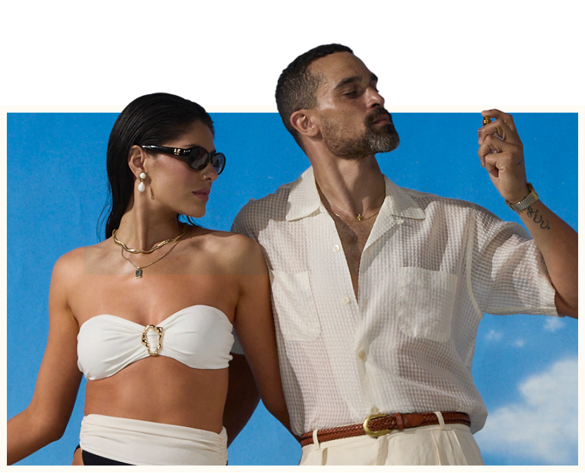 A woman in a white strapless top and sunglasses next to a man in a white shirt spraying fragrance, on a blue background.
