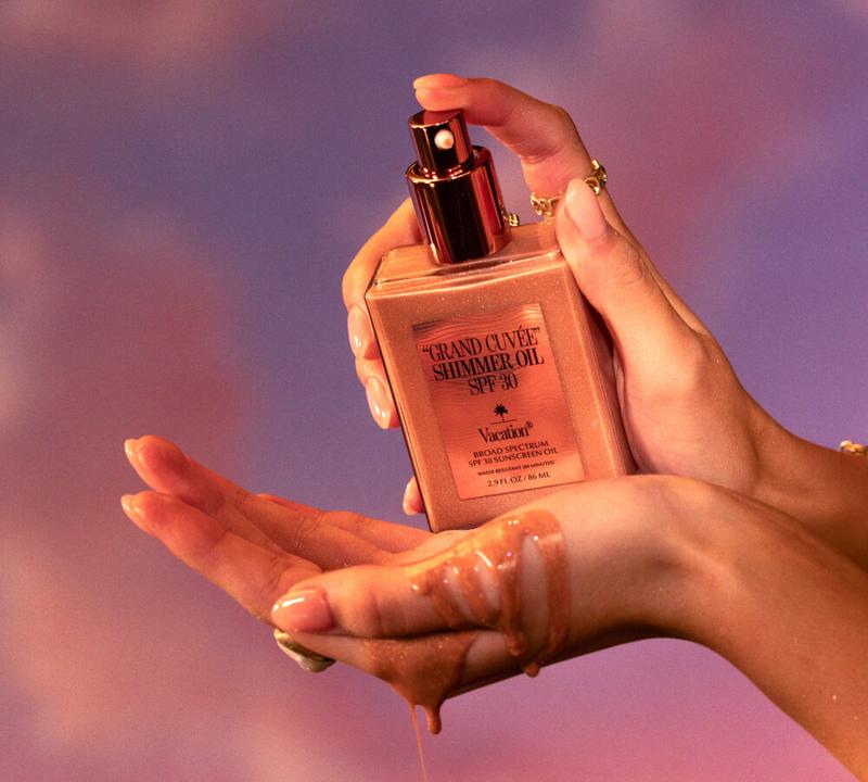 Hands dispensing Grand Cuvée Shimmer Oil SPF 30 from a square bottle, with the glistening oil dripping onto a palm.