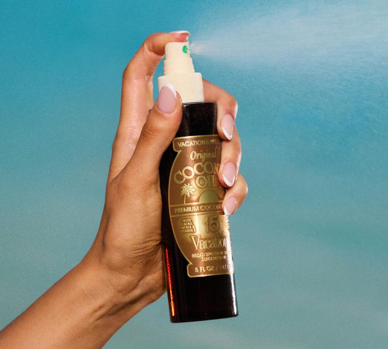 A hand sprays Vacation Coconut Oil SPF 15 from a bottle.