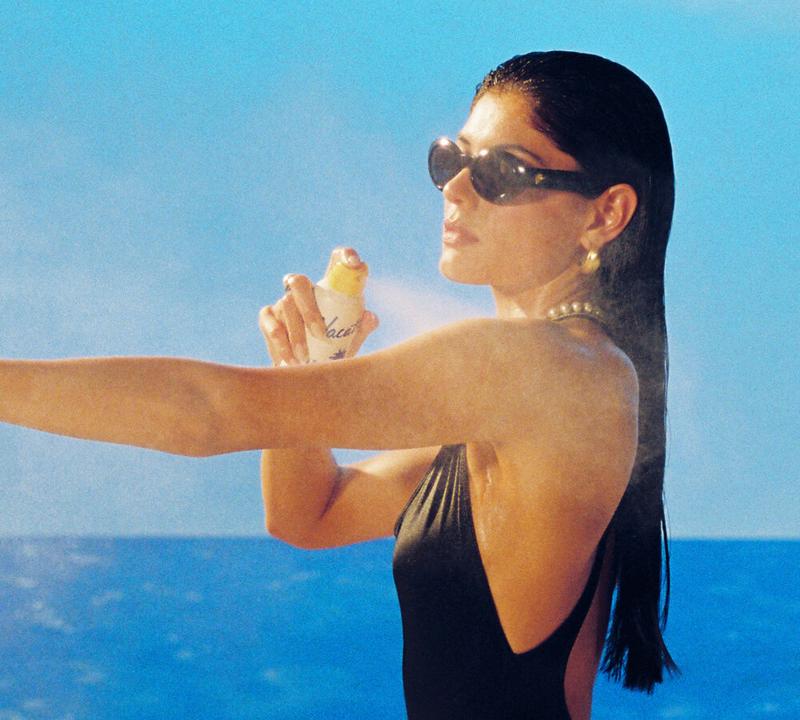 Woman in sunglasses and a black swimsuit spraying sunscreen on her arm.