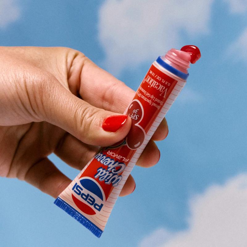 A hand with red nail polish holds a Pepsi Wild Cherry Flavor SPF 30 lip gel tube, with a dollop of red product emerging, against a blue sky.