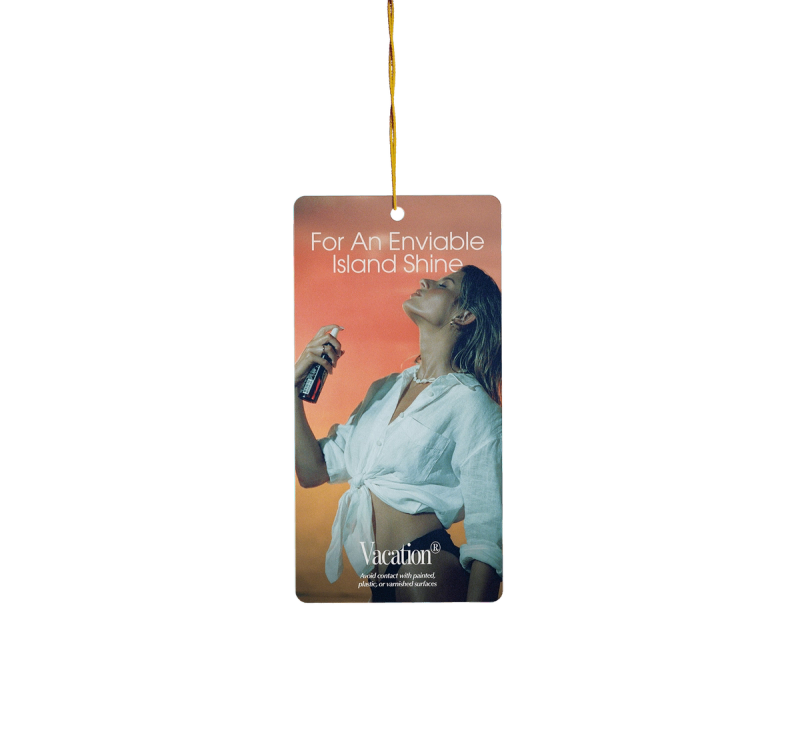 Rectangular air freshener featuring a woman spraying her face, with text "For An Enviable Island Shine" and "Vacation®".