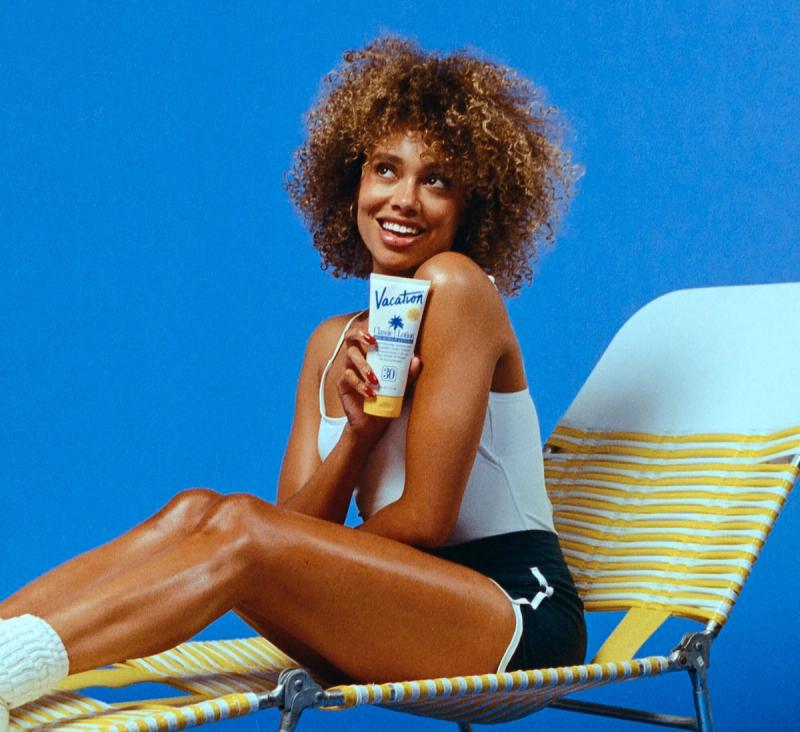 A smiling woman with curly hair sits in a beach chair, holding a tube of Vacation sunscreen.