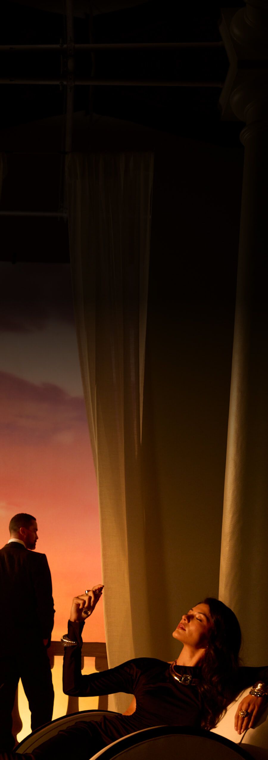 A woman in a dark dress and jewelry reclines, eyes closed, in warm light. To her left, a man in a suit looks out at a vibrant orange and pink sunset beyond columns and sheer curtains.