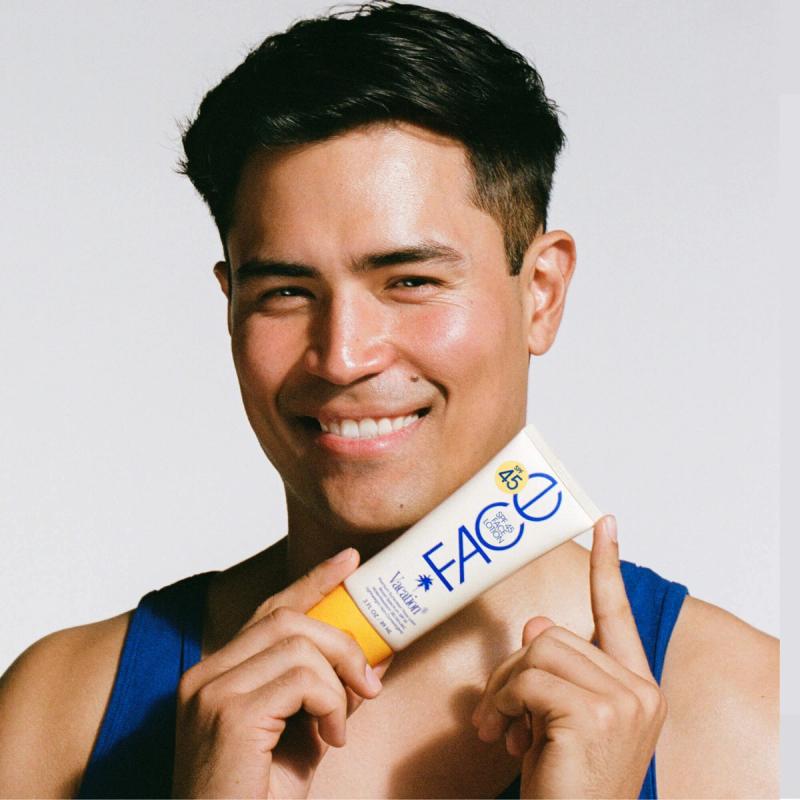 A smiling man in a blue tank top holds up a tube of Vacation FACE SPF 45 sunscreen.
