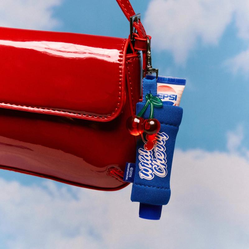 A glossy red purse with a blue keychain featuring red cherry charms and a Pepsi Wild Cherry lip balm, against a cloudy blue sky.