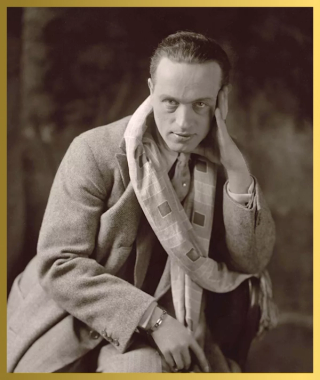 A sepia-toned portrait of a man in a tweed jacket and patterned scarf, looking at the camera with his hand on his temple.