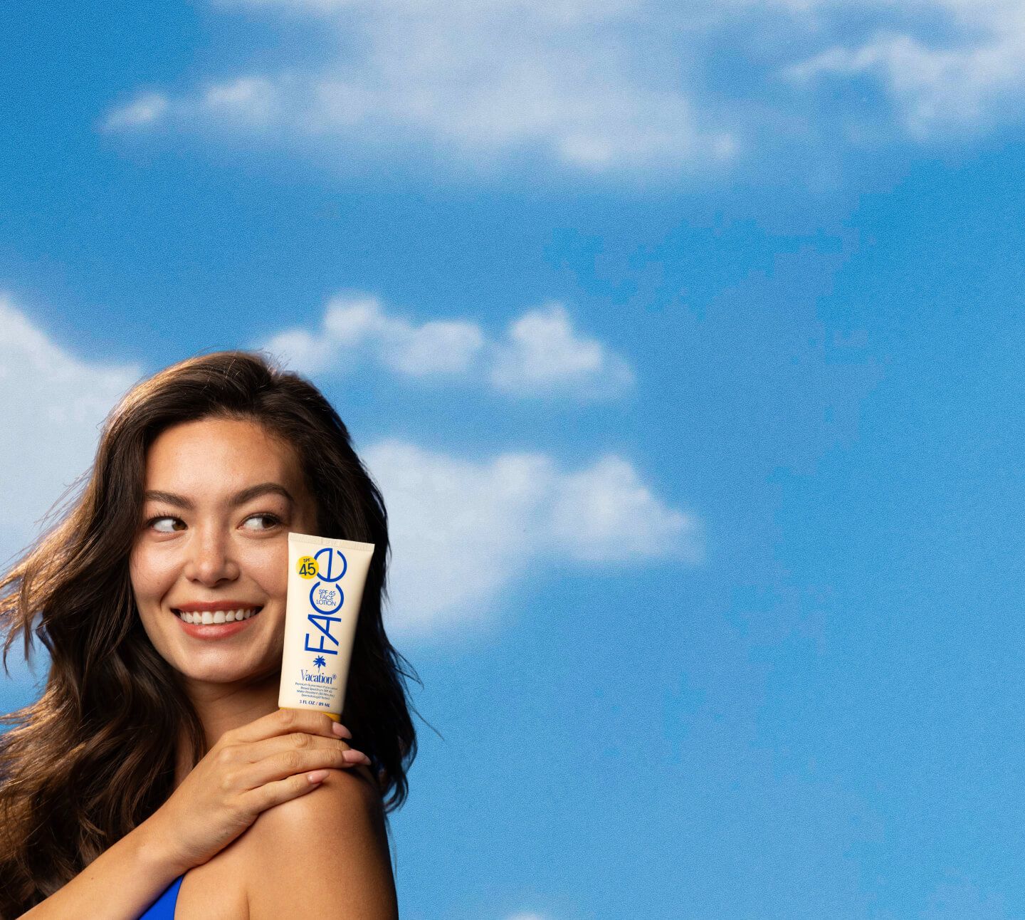 A smiling woman holds a tube of FACE SPF 45 sunscreen against a blue sky with clouds.
