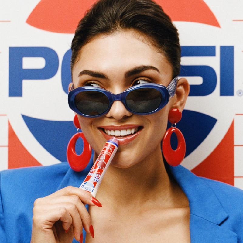 A smiling woman in blue sunglasses and red earrings holds a red and white lip gloss tube in her mouth, with a Pepsi logo in the background.