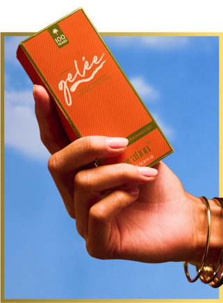 A hand holds an orange "Vacation" Gelée fragrance set box against a blue sky.