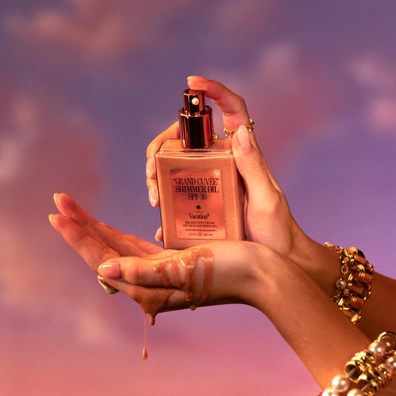 A person's hands hold a bottle of Grand Cuvée Shimmer Oil SPF 30, with the shimmering oil dripping onto one hand, against a purple and pink sky.