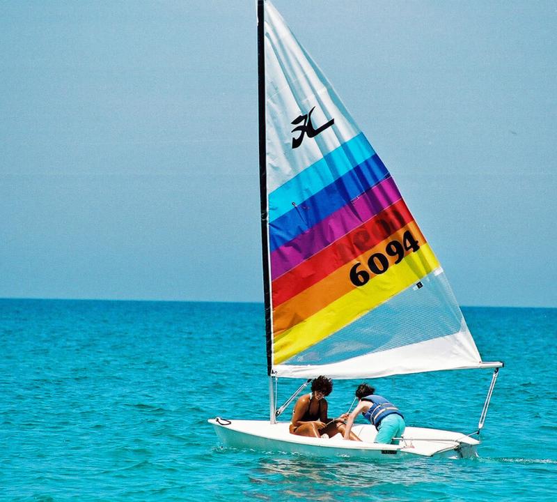 Two people on a sailboat with a rainbow-striped sail in the ocean.