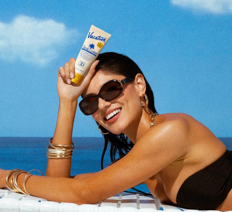 A smiling woman in sunglasses holds up a tube of Vacation sunscreen while lounging outdoors.