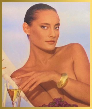 A woman with dark hair and a pearl earring reclines, topless, holding a glass of white wine next to purple grapes, against a blue sky background.